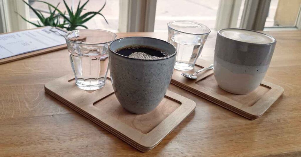 two ceramic mugs with cofee in one is a black coffee, the other is a flat white. They are on a little wonnde tray each with a glass of water. Discover where to find the best cofee in Seville in this article. 