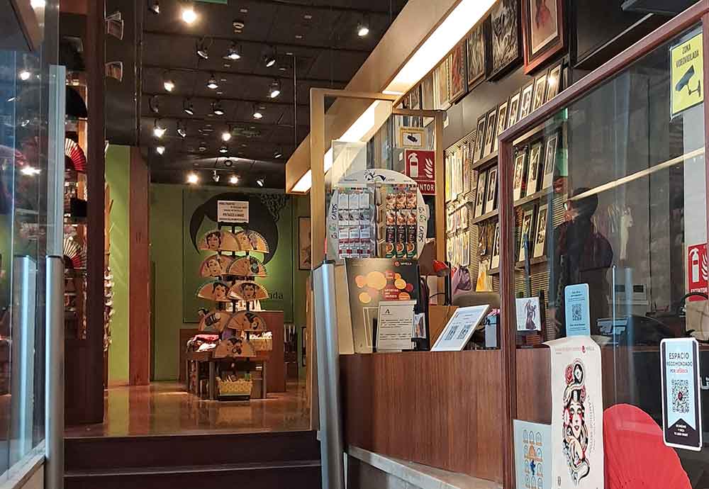 Museo del Baile Flamenco Front entrance of the Museo del Baile Flamenco in Seville, viewed from a narrow cobblestone street. Tall, elegant windows and red “Sevilla” banners hang above a dark doorway, while a small group of people stands nearby, hinting at the vibrant cultural experiences that await inside.