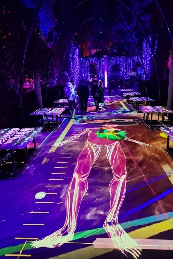An illuminated walkway at the Real Alcázar during the Naturaleza Encendida light show, with vibrant neon projections on the floor and glowing purple fairy lights creating a tunnel-like effect.