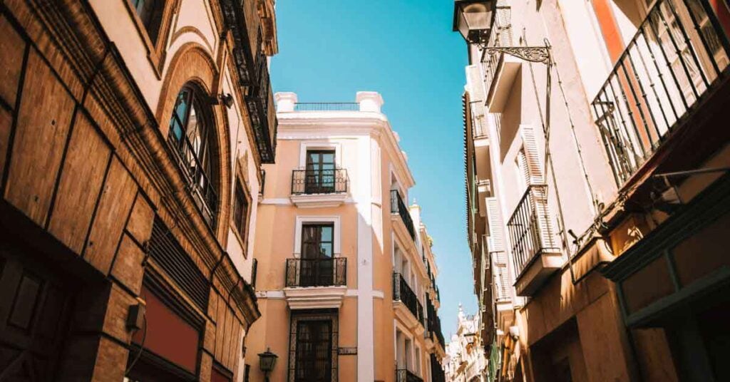 Sunny streets in September in Seville A narrow street in Seville, Spain, with traditional buildings and wrought-iron balconies lit by warm September sunshine under a clear blue sky — a typical scene when exploring the best things to do in Seville in September.