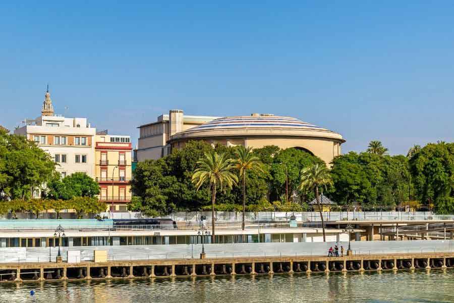 Teatro de la Maestranza View across the Guadalquivir River in Seville, showing palm trees, greenery, and the circular dome of the Teatro de la Maestranza. Residential buildings and the Giralda tower rise in the background under a clear blue sky.